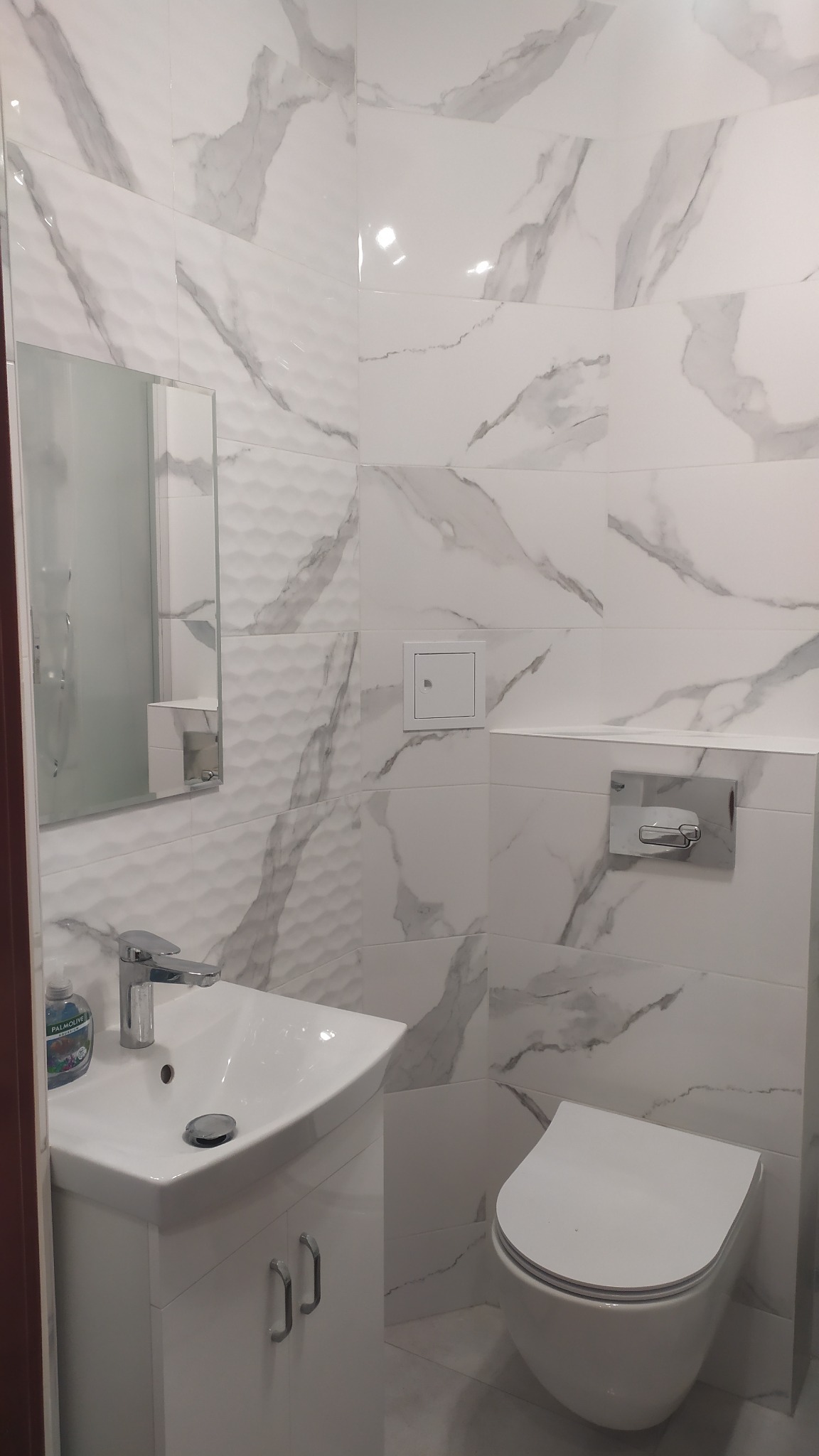 Nowoczesna łazienka z białymi płytkami imitującymi marmur, wisząca toaleta, umywalka z szafką i lustro z oświetleniem punktowym.