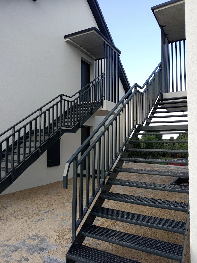 Zewnętrzne, dwubiegowe schody metalowe z ażurowymi ściankami i balustradami, ocynkowane i malowane proszkowo na kolor RAL 7016, prowadzące do dwóch wejść.