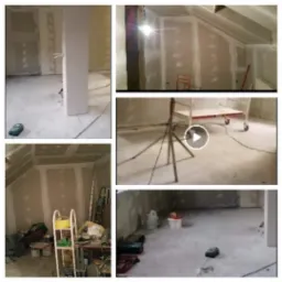 Wnętrze w trakcie wykańczania: ściany pokryte płytami gipsowo-kartonowymi, widoczne szpachlowanie łączeń, drabina, narzędzia i prowizoryczne oświetlenie. Na podłodze widoczne ślady zaprawy.