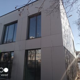 Chufu usługi budowlane - Montaż elewacji budynku z szarych paneli kompozytowych, dwóch pracowników na rusztowaniu, widoczne okna z odbiciem drzew, boczny widok budynku w słoneczny dzień.