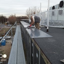 Chufu usługi budowlane - Pracownik w czerwonych rękawicach montuje metalowe panele na dachu budynku przemysłowego, widoczne rusztowanie, izolacja, wentylatory i otoczenie miejskie.