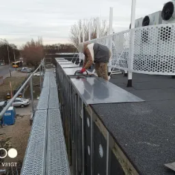 Pracownik w czerwonych rękawicach montuje metalowe panele na dachu budynku przemysłowego, widoczne rusztowanie, izolacja, wentylatory i otoczenie miejskie.