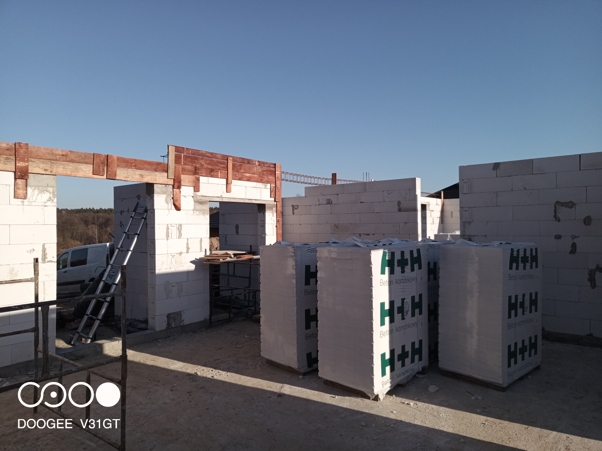 Wznoszenie ścian domu z betonu komórkowego H+H, widoczne drewniane nadproża okienne i drzwiowe, drabina oparta o ścianę, w tle las i błękitne niebo.