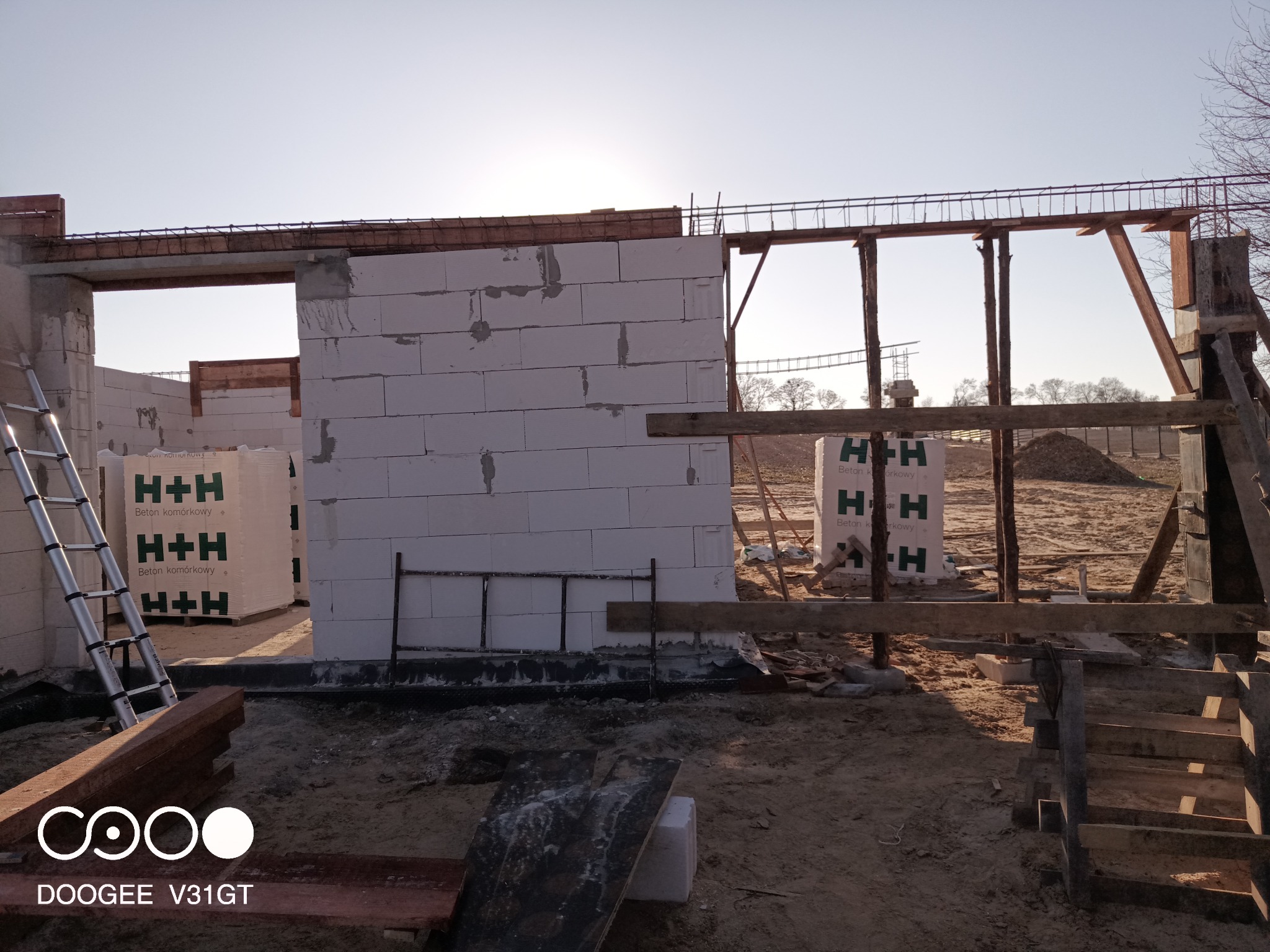 Wznoszenie ścian z betonu komórkowego na budowie w Łodzi, widoczne palety z materiałem H+H, szalunki i zbrojenia.