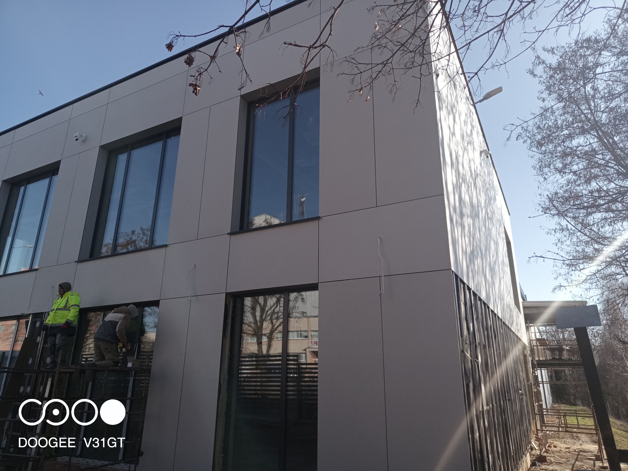 Montaż elewacji budynku z szarych paneli kompozytowych, dwóch pracowników na rusztowaniu, widoczne okna z odbiciem drzew, boczny widok budynku w słoneczny dzień.