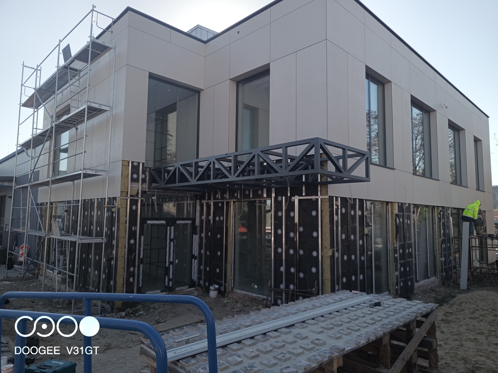 Budynek w trakcie ocieplania elewacji, widoczne rusztowania, warstwa izolacyjna i częściowo zamontowane panele elewacyjne. Stalowa konstrukcja nad wejściem. Robotnik w jaskrawej kurtce w tle.