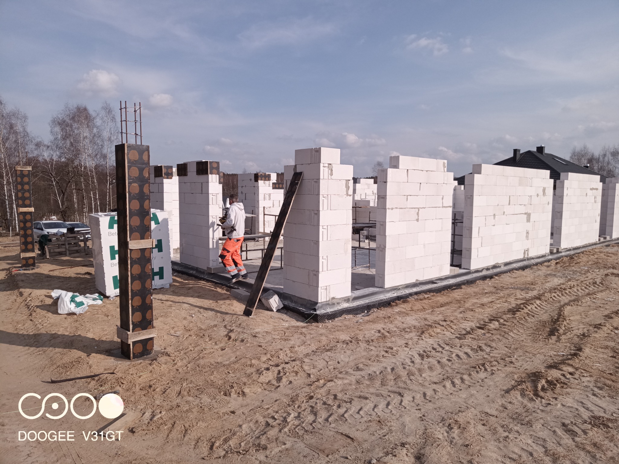 Wznoszenie ścian domu z białych bloczków, widoczne zbrojenia słupów, pracownik w pomarańczowych spodniach na rusztowaniu, teren budowy z piaskiem i śladami opon