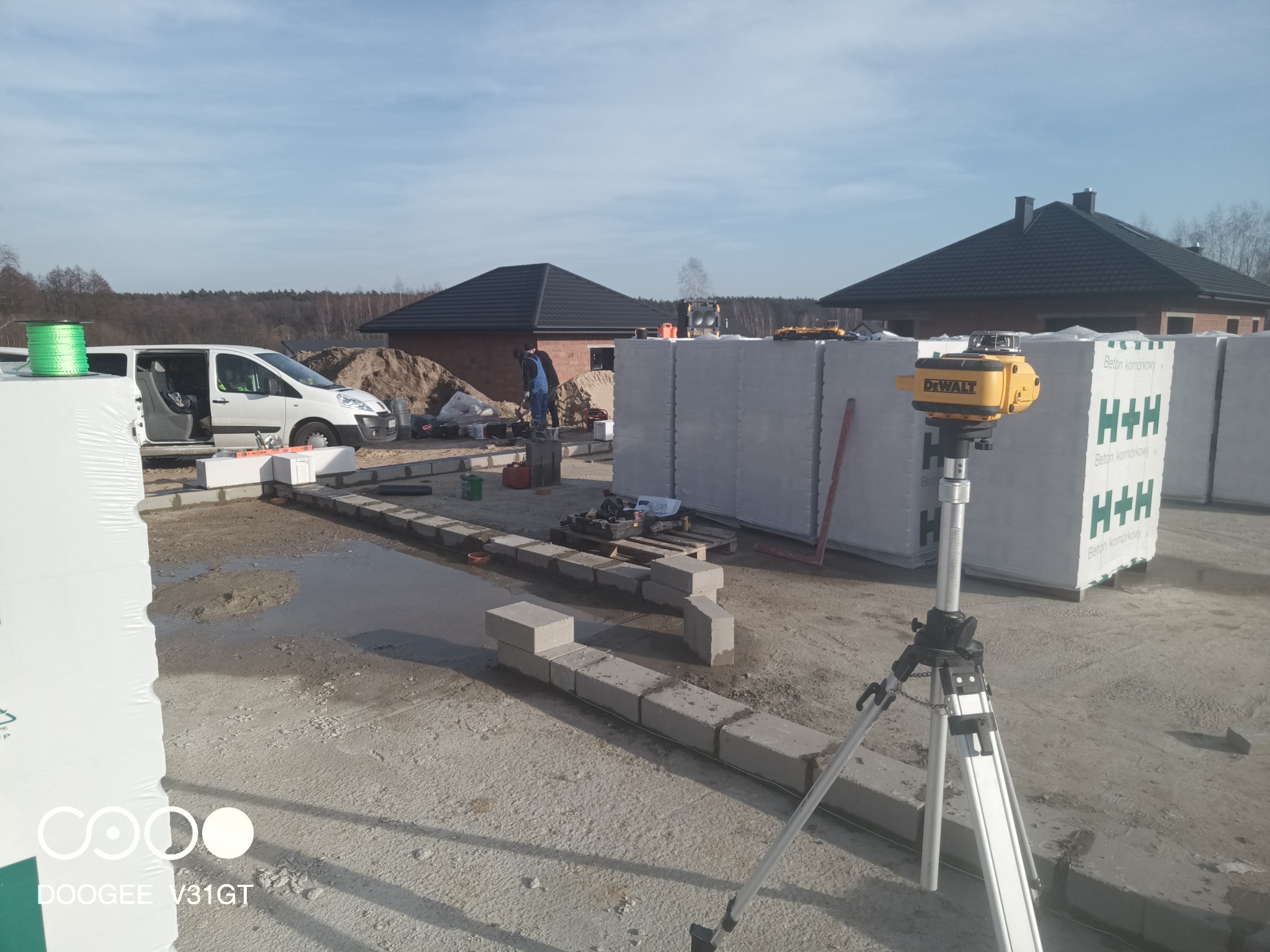 Wznoszenie ścian fundamentowych z bloczków betonowych H+H, widoczny poziomica laserowa DeWalt na statywie, palety z bloczkami, oraz pracownicy budowlani w tle.