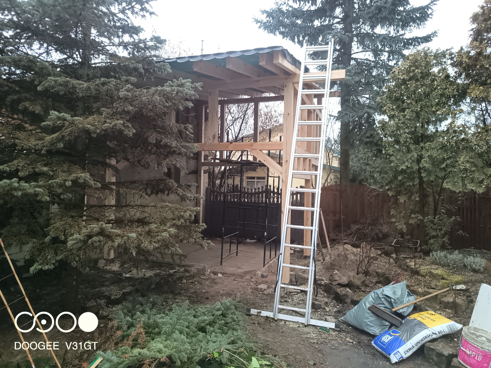 Konstrukcja drewniana z częściowo położonym dachem z blachy, widoczna drabina aluminiowa, worki z ziemią i narzędzia ogrodnicze w otoczeniu drzew i krzewów.