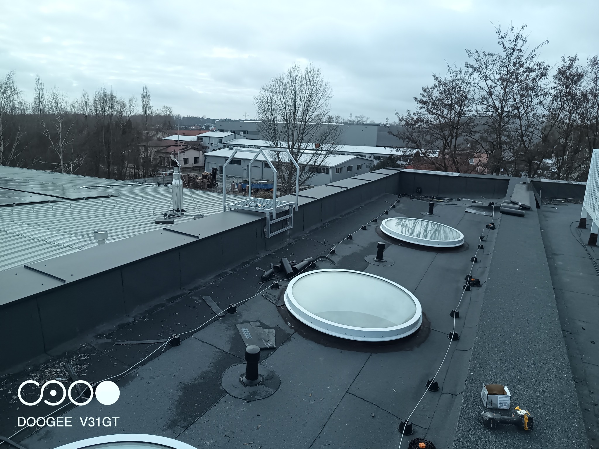 Płaski dach pokryty papą z trzema świetlikami w kształcie okręgów, widoczne narzędzia i materiały budowlane oraz otoczenie z budynkami i drzewami w tle.