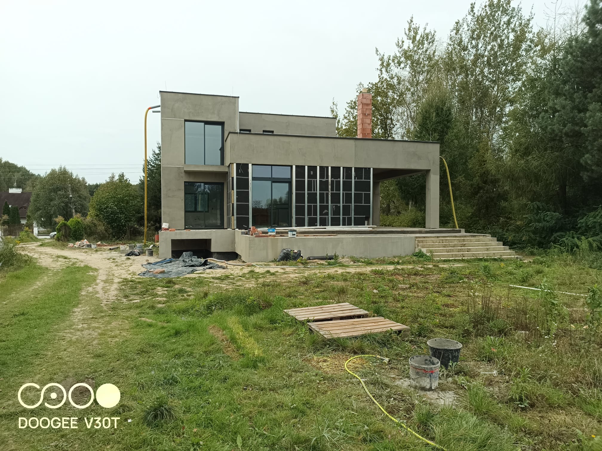 Surowy, nowoczesny dom jednorodzinny w trakcie budowy, z widocznym ociepleniem ścian i dużymi oknami, otoczony niezagospodarowanym terenem z paletami i wiadrami budowlanymi.