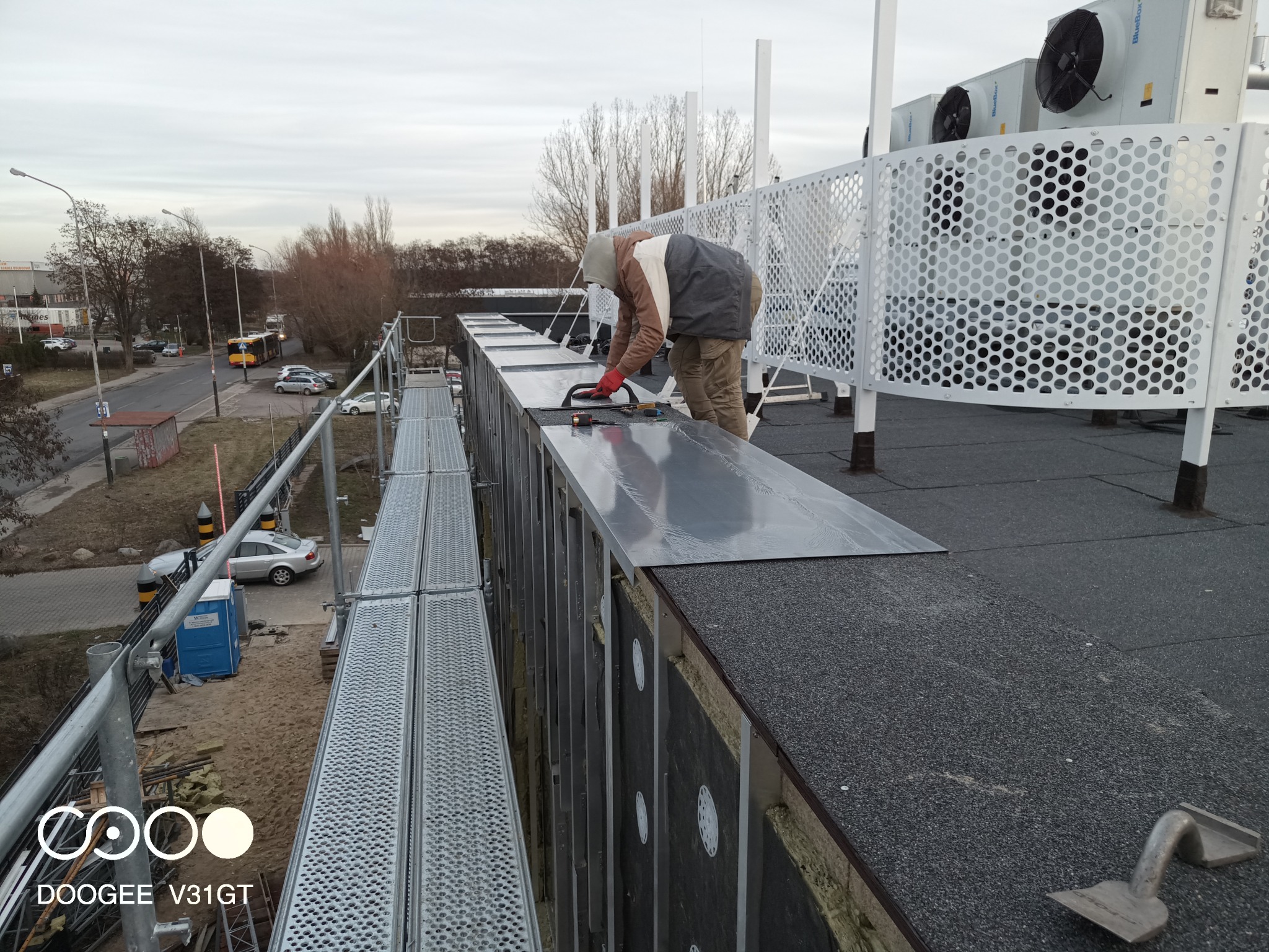 Pracownik w czerwonych rękawicach montuje metalowe panele na dachu budynku przemysłowego, widoczne rusztowanie, izolacja, wentylatory i otoczenie miejskie.