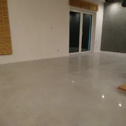 Wykończone wnętrze z jasną, połyskującą podłogą z płytek, drewnianymi żaluzjami i ścianą w odcieniach szarości, widoczne fragmenty schodów.