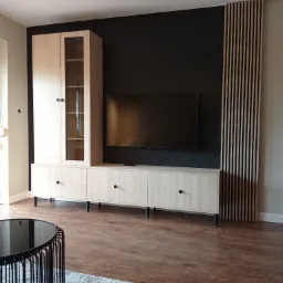 Jasna szafka RTV z witryną, czarne tło ściany z telewizorem, drewniane lamele pionowe, parkiet i fragment stolika kawowego.
