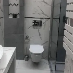 Nowoczesna łazienka z białymi i szarymi płytkami imitującymi marmur, wisząca toaleta z chromowanym przyciskiem, szklana kabina prysznicowa i dekoracyjna nisza z rośliną.