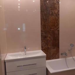 Perfect Job Sp. z o.o. - Wykończona łazienka z beżowymi płytkami, akcentem z brązowego marmuru, białą szafką pod umywalką z dwiema szufladami i wanną z prysznicem.