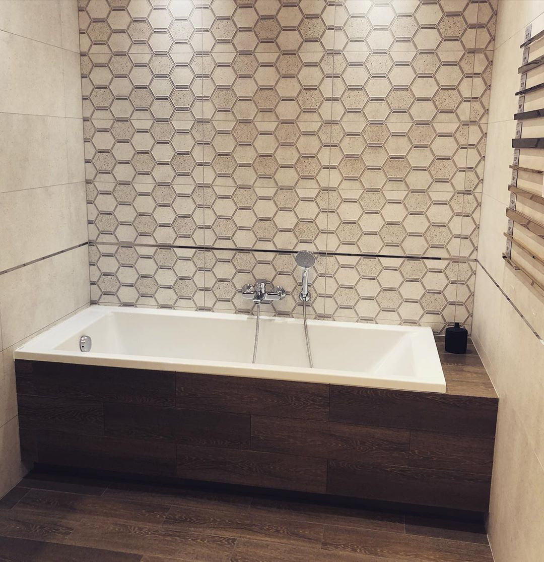 Biała wanna z obudową z paneli imitujących drewno, na tle ściany wyłożonej heksagonalnymi płytkami w odcieniach beżu i szarości, z chromowaną baterią i słuchawką prysznicową.