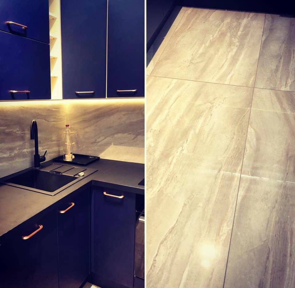 Nowoczesna kuchnia w Ząbkach: ciemnoniebieskie szafki z miedzianymi uchwytami, czarny zlew i armatura, marmurowy blat oraz podłoga wyłożona płytkami imitującymi marmur.