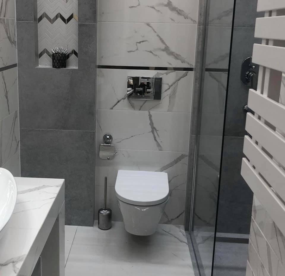 Nowoczesna łazienka z białymi i szarymi płytkami imitującymi marmur, wisząca toaleta z chromowanym przyciskiem, szklana kabina prysznicowa i dekoracyjna nisza z rośliną.