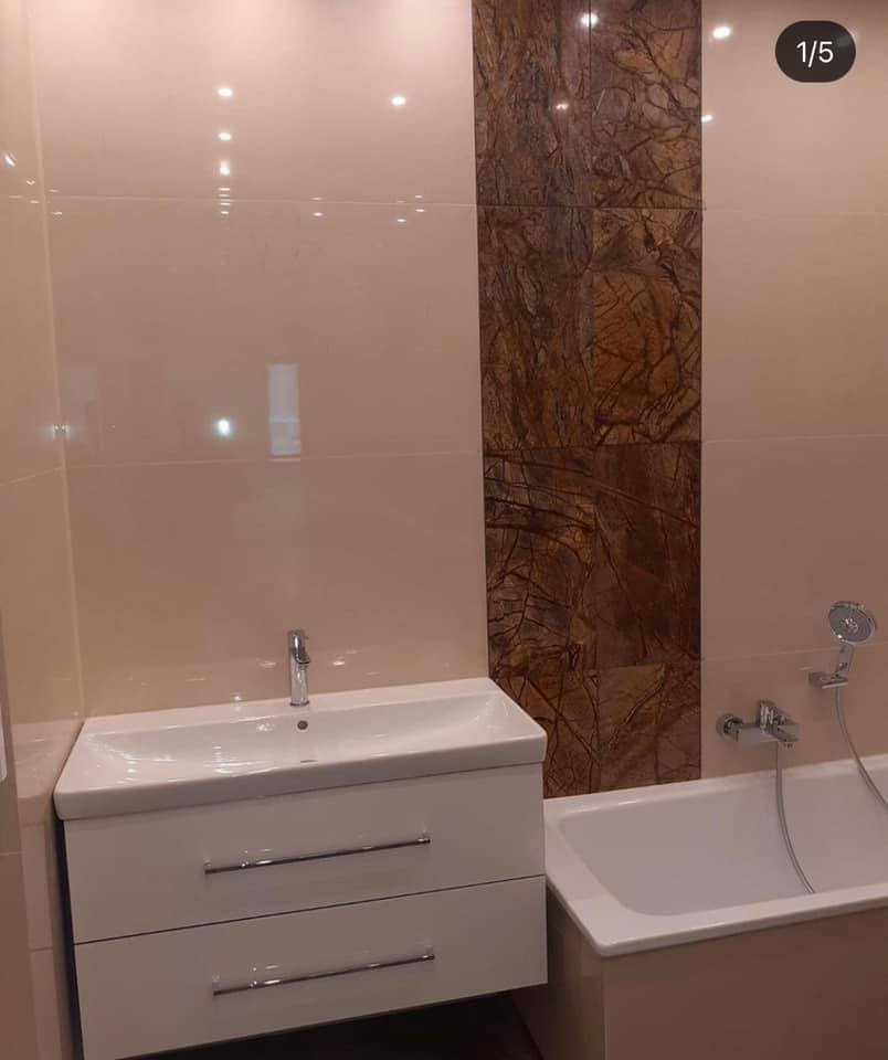 Wykończona łazienka z beżowymi płytkami, akcentem z brązowego marmuru, białą szafką pod umywalką z dwiema szufladami i wanną z prysznicem.