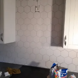 Układanie heksagonalnych płytek na ścianie w kuchni, widoczna puszka elektryczna bez osprzętu, białe szafki kuchenne po bokach, na blacie worek z zaprawą klejową Mapei Ultracolor Plus.