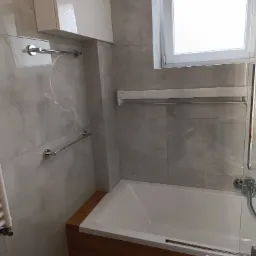 Nowoczesna łazienka z wanną obudowaną drewnem, szare płytki imitujące marmur, grzejnik drabinkowy i szklany parawan nawannowy.