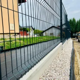 Nowoczesne, czarne ogrodzenie panelowe z podmurówką betonową i podsypką z białego grysu, widok z boku na tle zabudowań i drzew.