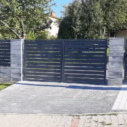 Nowoczesna, pozioma brama wjazdowa i furtka w kolorze antracytowym, z murowanymi słupkami z szarej kostki, na tle zieleni drzew i domu. Podjazd wyłożony szarą kostką brukową z elementami białej...