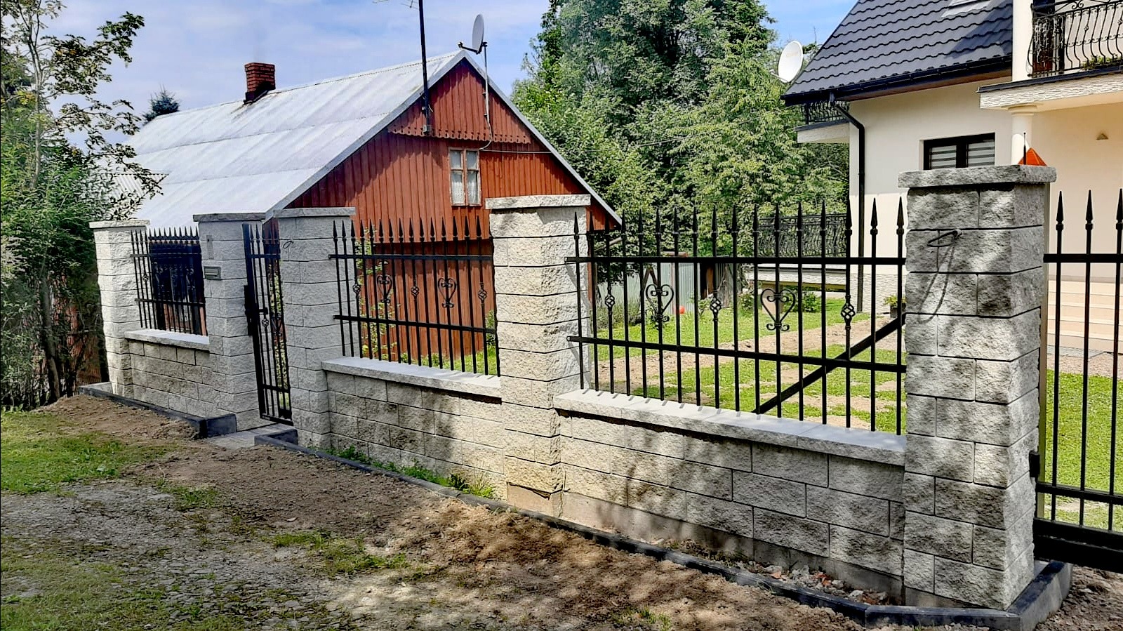 Nowoczesne ogrodzenie z czarnych, kutych przęseł między słupkami z szarej kostki łupanej, z furtką i podmurówką, na tle dwóch domów i zieleni.