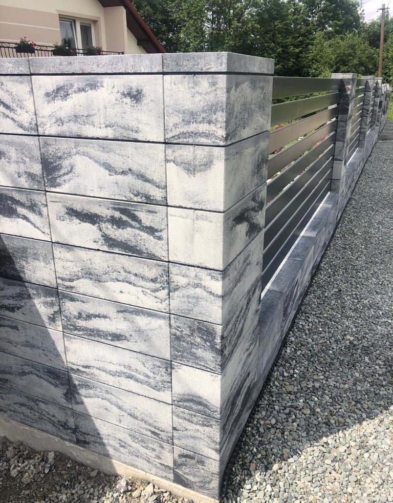 Nowoczesne ogrodzenie z betonowych bloczków o wzorze imitującym kamień i poziomych metalowych przęseł, otoczone szarym żwirem