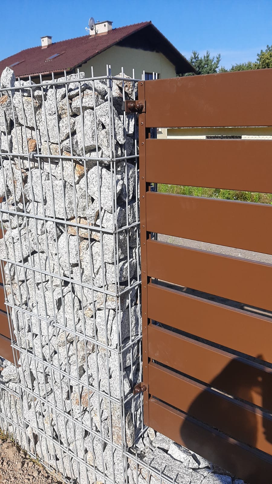 Fragment ogrodzenia: gabion z jasnymi kamieniami połączony z brązowymi, poziomymi panelami drewnopodobnymi, widoczny cień osoby, w tle dom jednorodzinny.