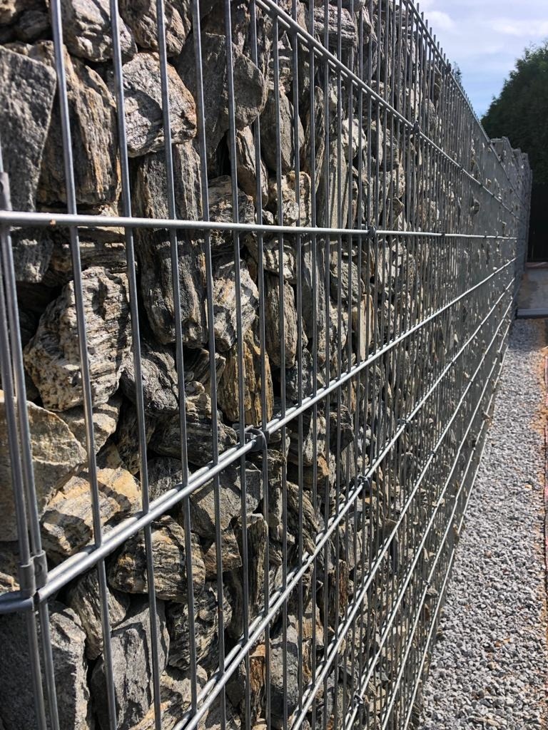 Długi mur z gabionów wypełnionych szaro-brązowymi kamieniami, widoczny z boku, z drobnym żwirem u podstawy i zielenią w tle.