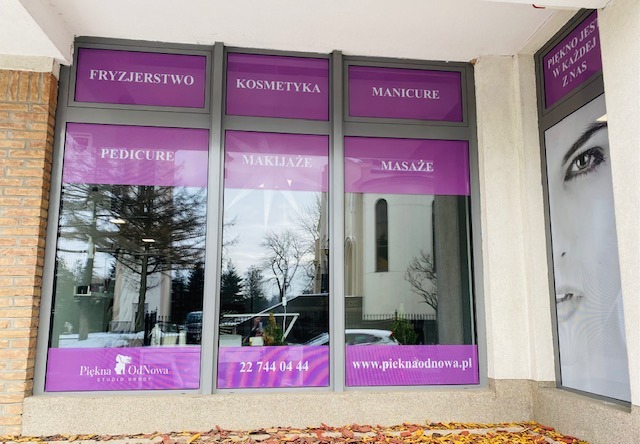 Fasada budynku salonu urody z dużymi oknami, na których widnieją nazwy usług: Fryzjerstwo, Kosmetyka, Manicure, Pedicure, Makijaże, Masaże oraz logo firmy Piekna OdNowa. W szybach odbija się...