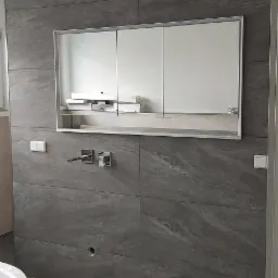 Szara łazienka z płytkami imitującymi kamień, nowoczesna armatura, szafka z lustrem i biała wanna. Na podłodze leży szary dywanik.