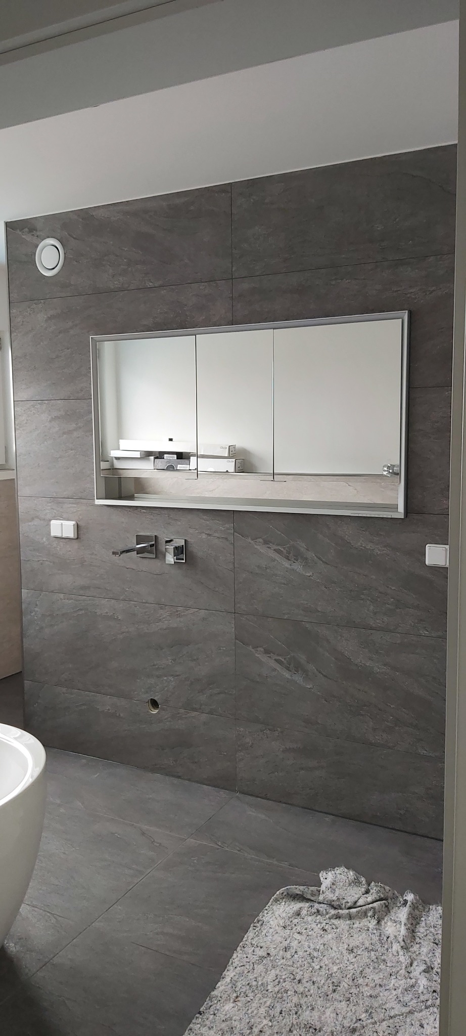 Szara łazienka z płytkami imitującymi kamień, nowoczesna armatura, szafka z lustrem i biała wanna. Na podłodze leży szary dywanik.