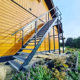 Zewnętrzne schody metalowe z kratowymi stopniami, prowadzące do drewnianego budynku w Żarach, otoczone rabatami kwiatowymi.
