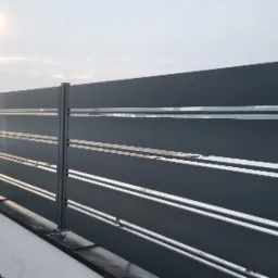 Nowoczesne, poziome ogrodzenie panelowe w kolorze grafitowym z metalowymi poprzeczkami, zamontowane na niskim, białym murku, oświetlone lampą uliczną w tle.