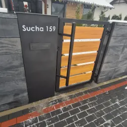 Nowoczesna brama wjazdowa z poziomymi drewnianymi panelami, część muru z szarej kostki z numerem domu 'Sucha 159' i skrzynką na listy, kostka brukowa z resztkami śniegu, dom z dekoracjami...