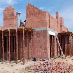 Wznoszenie ścian z czerwonej cegły na budowie domu, widoczne drewniane stemple podtrzymujące strop oraz stos potłuczonych cegieł na ziemi.