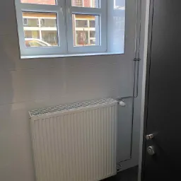 Biały grzejnik panelowy zamontowany na ścianie wyłożonej jasnymi płytkami, z widocznymi rurami doprowadzającymi, termostatem i oknem w tle, w pomieszczeniu z ciemną podłogą.