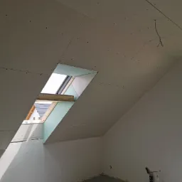 Poddasze w trakcie wykańczania płytami kartonowo-gipsowymi, widoczne fragmenty konstrukcji okna dachowego i wystające przewody elektryczne.