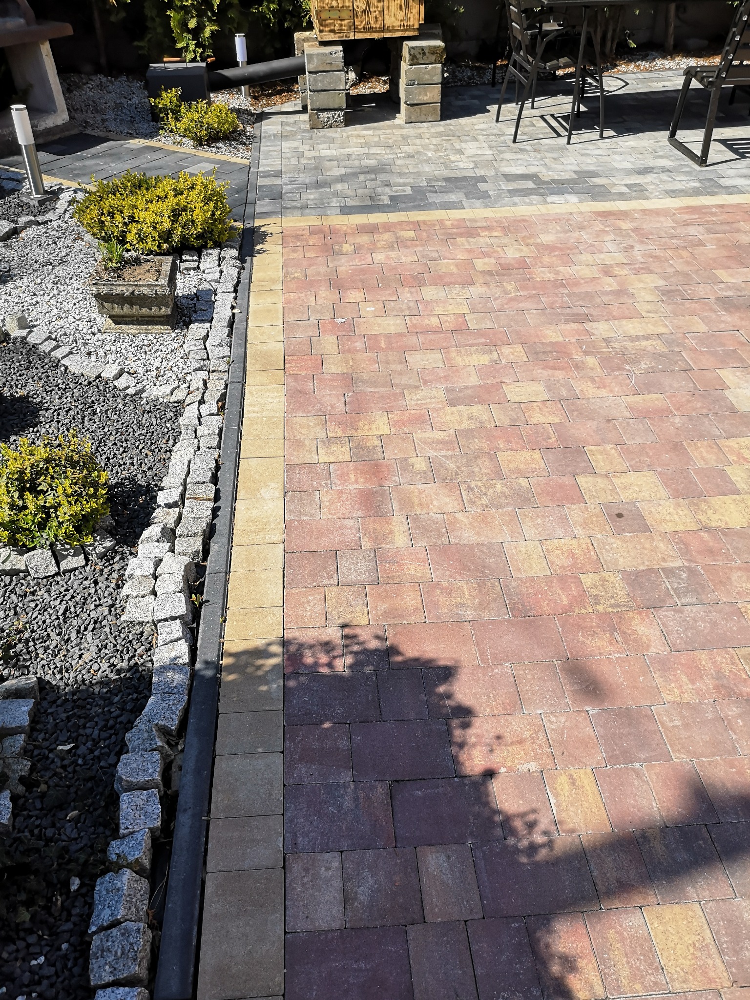 Ułożona nawierzchnia z kostki brukowej w odcieniach czerwieni i beżu, oddzielona obrzeżem od rabaty z szarym grysem i roślinnością. W tle widoczne meble ogrodowe.