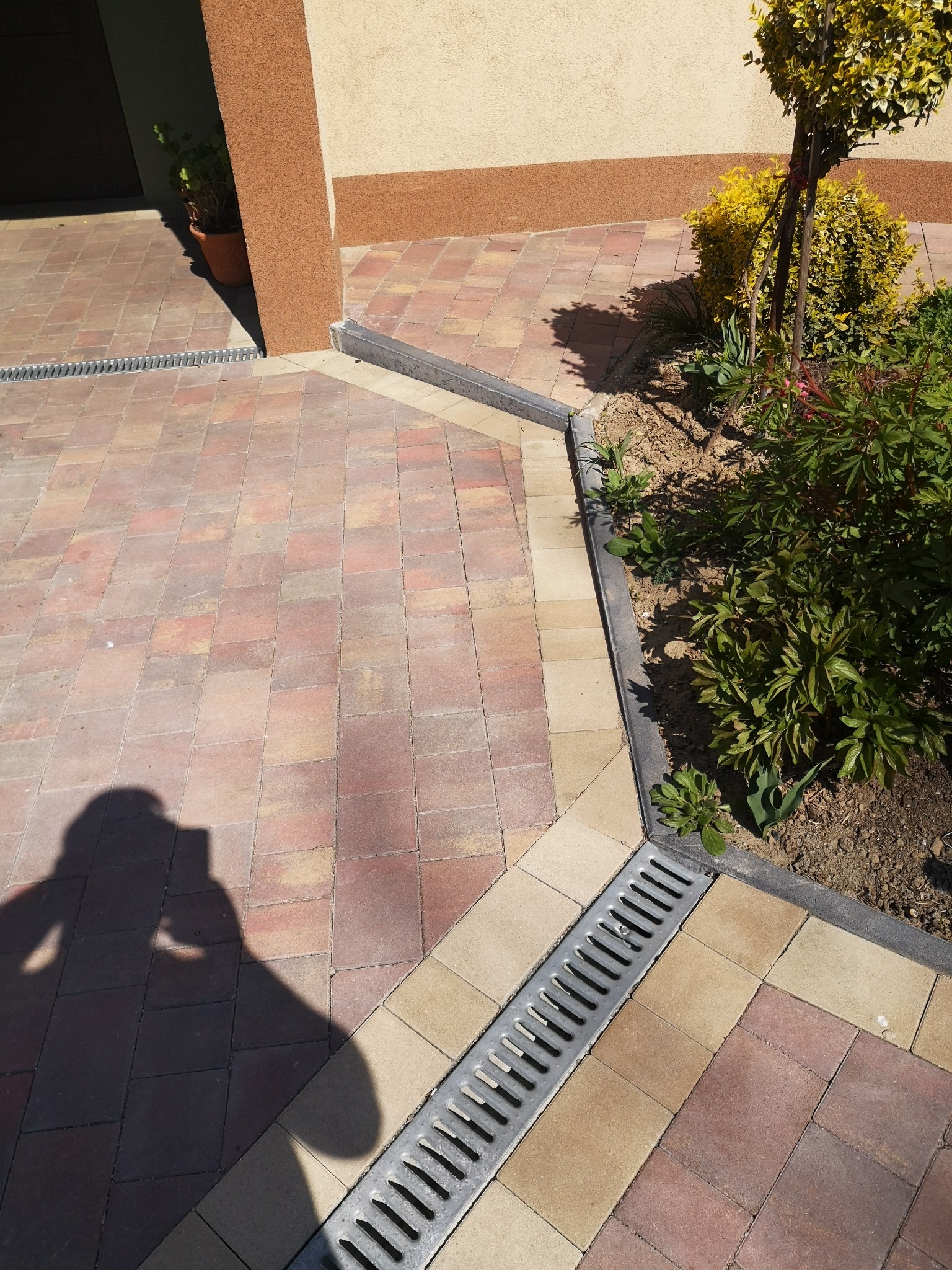 Nowo ułożona, wielobarwna kostka brukowa z odpływem liniowym, obrzeżami i zielenią w tle, cień osoby robiącej zdjęcie.