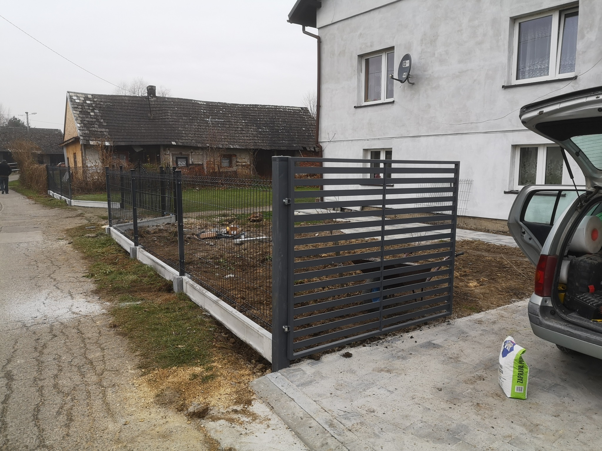 Nowoczesne, grafitowe ogrodzenie panelowe z podmurówką prefabrykowaną w trakcie montażu, widok na posesję z domem i otwarty bagażnik samochodu z narzędziami.