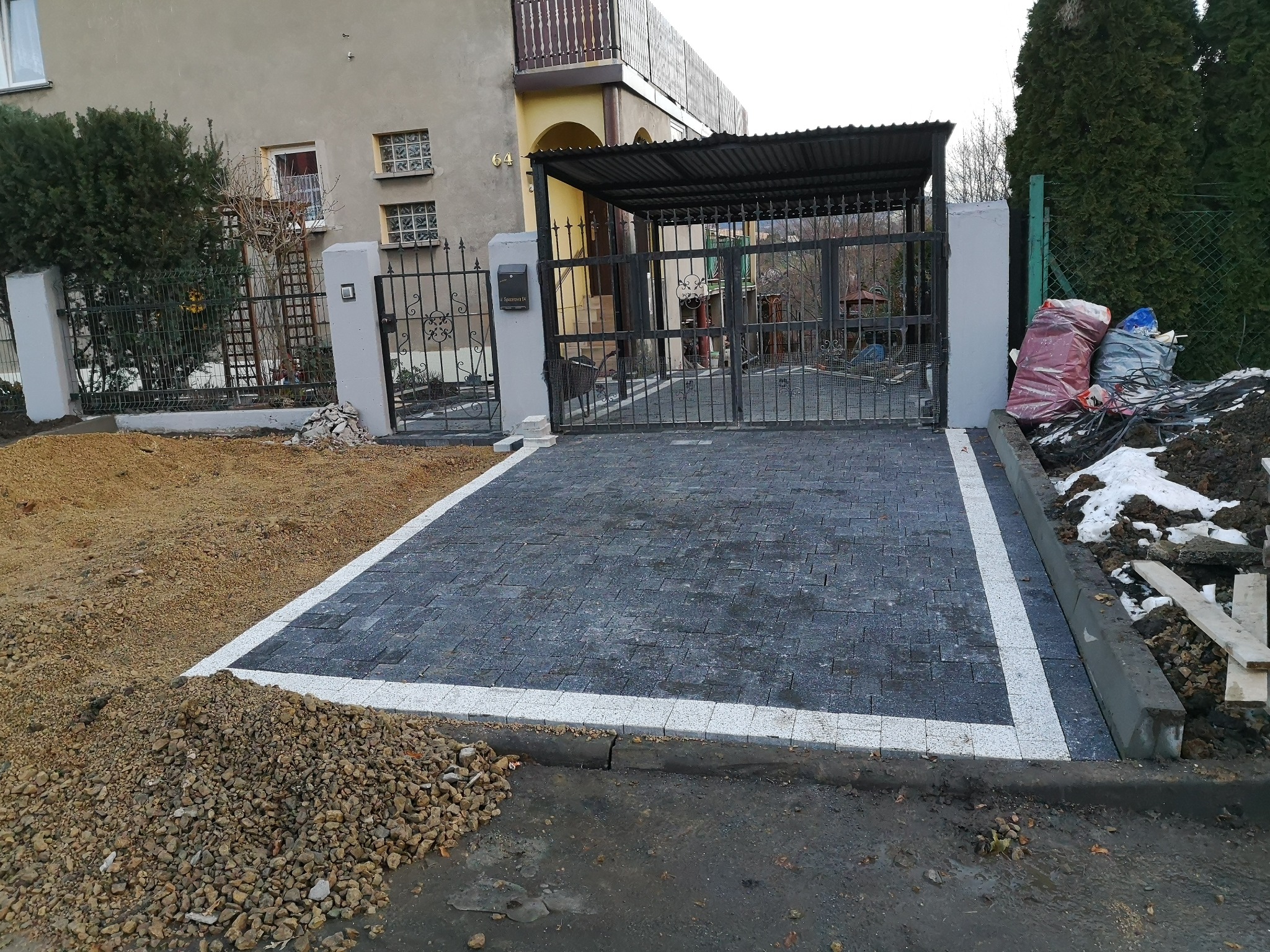 Ukończony podjazd z kostki brukowej w Bielsko-Białej, z wyraźnym obramowaniem z jasnej kostki, prowadzący do metalowej bramy wjazdowej pod zadaszeniem.