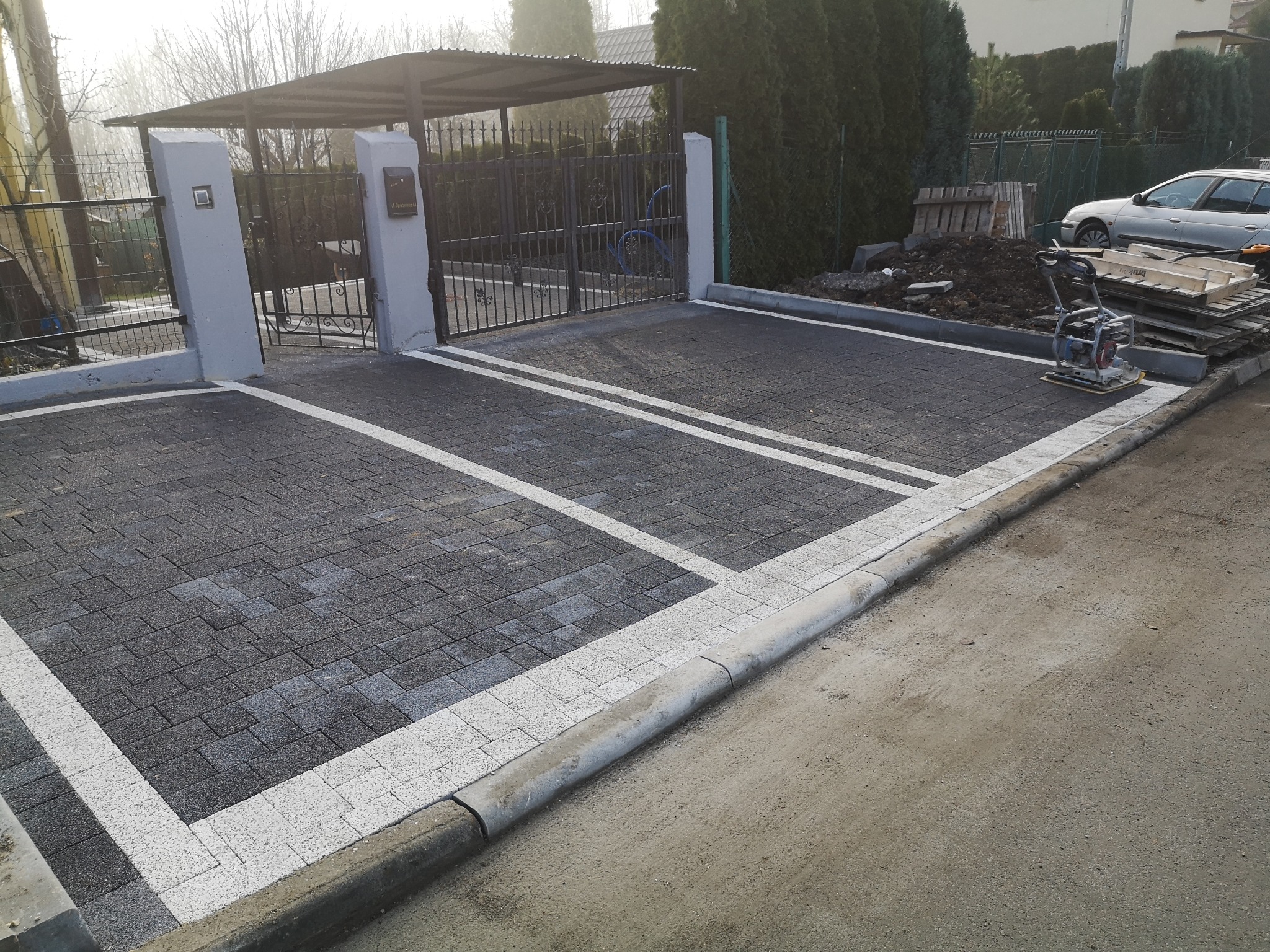 Ułożona, szara kostka brukowa z białymi pasami wyznaczającymi miejsca parkingowe, widoczna zagęszczarka wibracyjna, brama wjazdowa, samochód w tle.