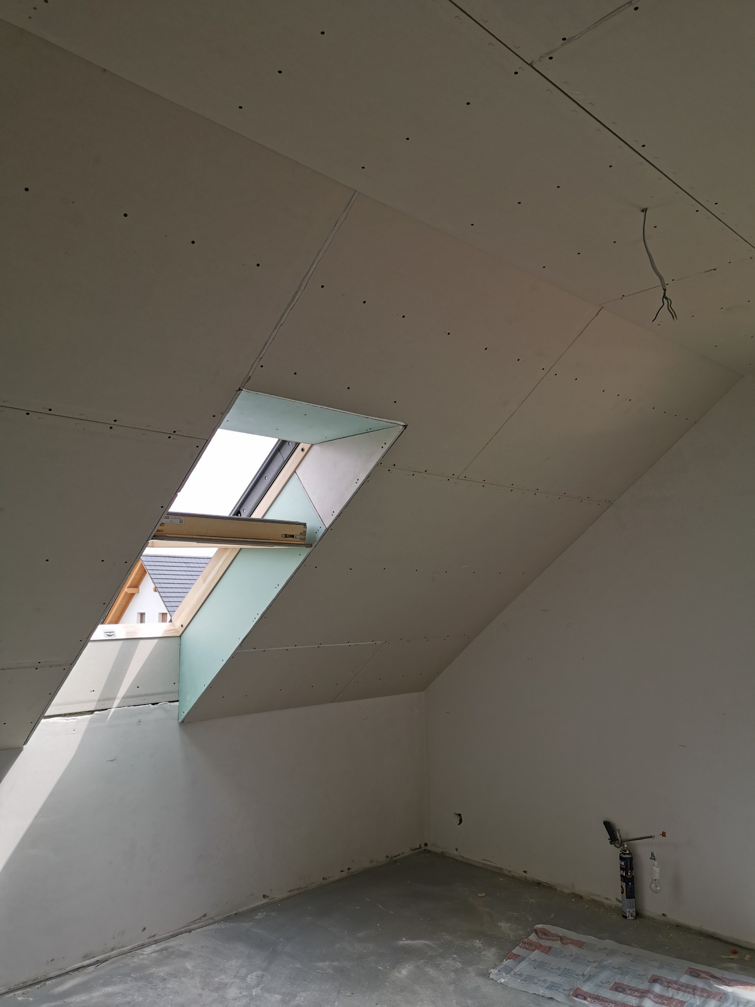 Poddasze w trakcie wykańczania płytami kartonowo-gipsowymi, widoczne fragmenty konstrukcji okna dachowego i wystające przewody elektryczne.