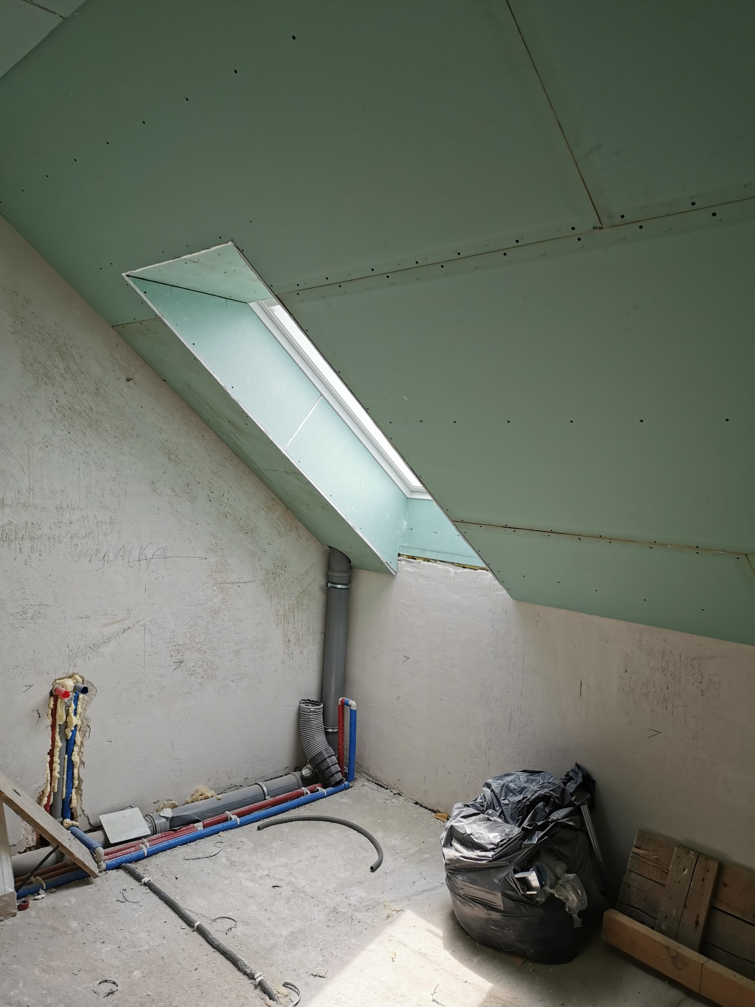 Poddasze w trakcie wykańczania: widoczne płyty kartonowo-gipsowe na skosach, okno dachowe, instalacja wodno-kanalizacyjna i worek z odpadami budowlanymi.