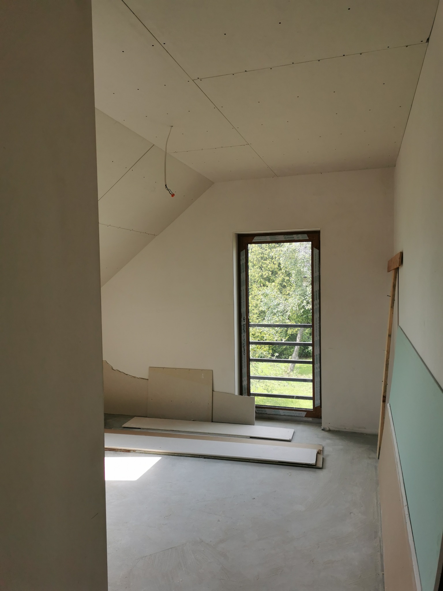 Poddasze w trakcie wykańczania płytami kartonowo-gipsowymi, widoczne łączenia na suficie, okno balkonowe z widokiem na zieleń oraz przygotowane płyty na podłodze.