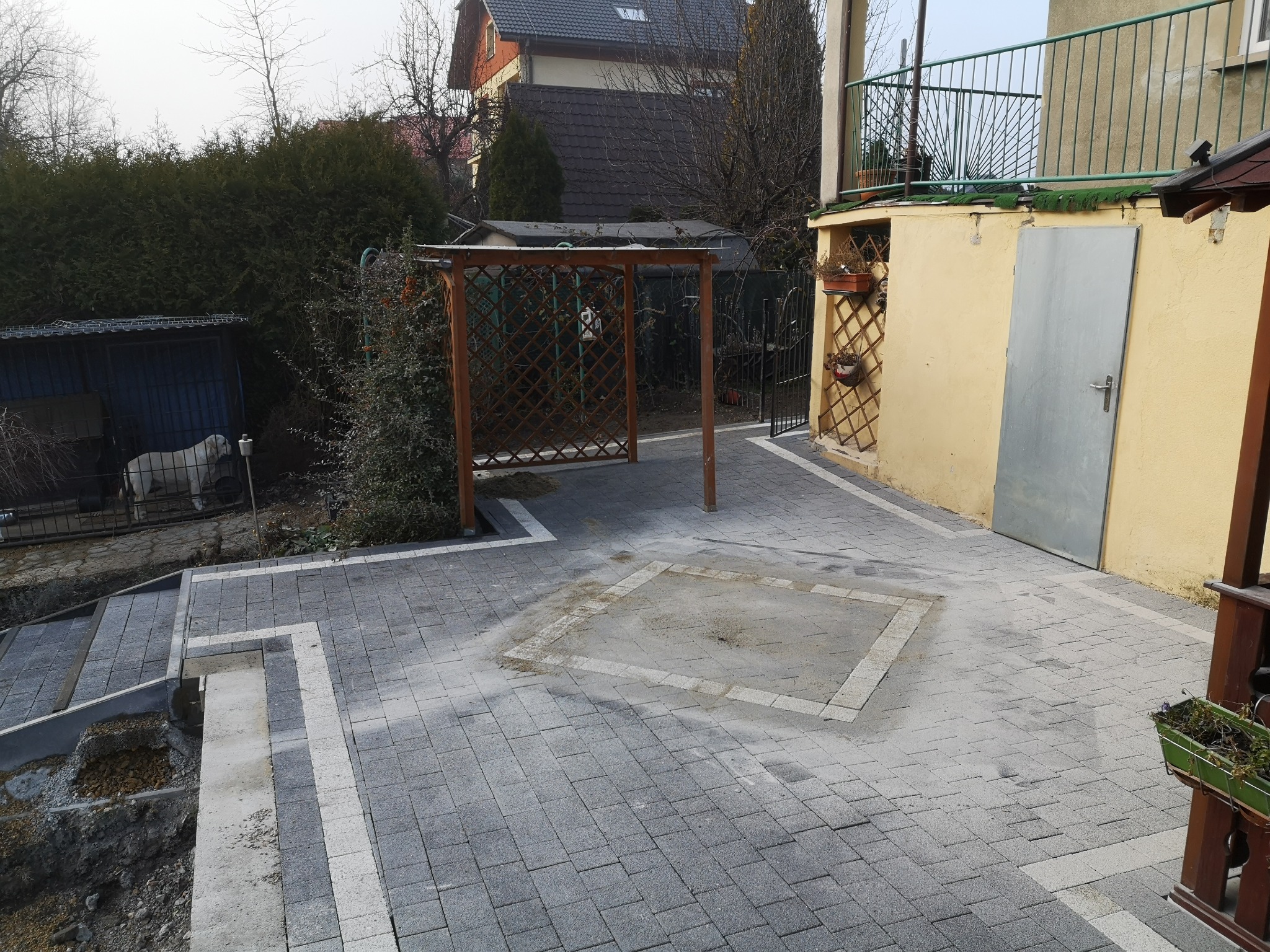 Ułożona szara kostka brukowa na podwórku z drewnianą pergolą i schodami, widoczny fragment budynku z żółtą elewacją i balkonem, oraz buda dla psa z białym psem w środku.